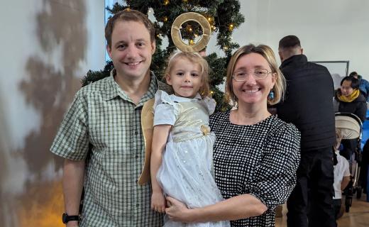 A family photo with Geoffrey Hayward wearing a green plaid shirt, his youngest child Lizzie in a white angel costume with golden wings and a halo, and Ewa Hayward wearing a black and white patterned dress. They are standing in front of a Christmas tree, smiling at the camera, with other people and holiday decorations blurred in the background. The photo was taken after Lizzie's school nativity show.