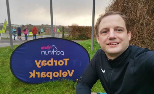 Geoffrey Hayward seated next to the Velopark Torbay parkrun sign, capturing a selfie. He is clad in a black sports outfit and appears content. The backdrop features a cloudy sky with other participants and volunteers visible in a park environment.