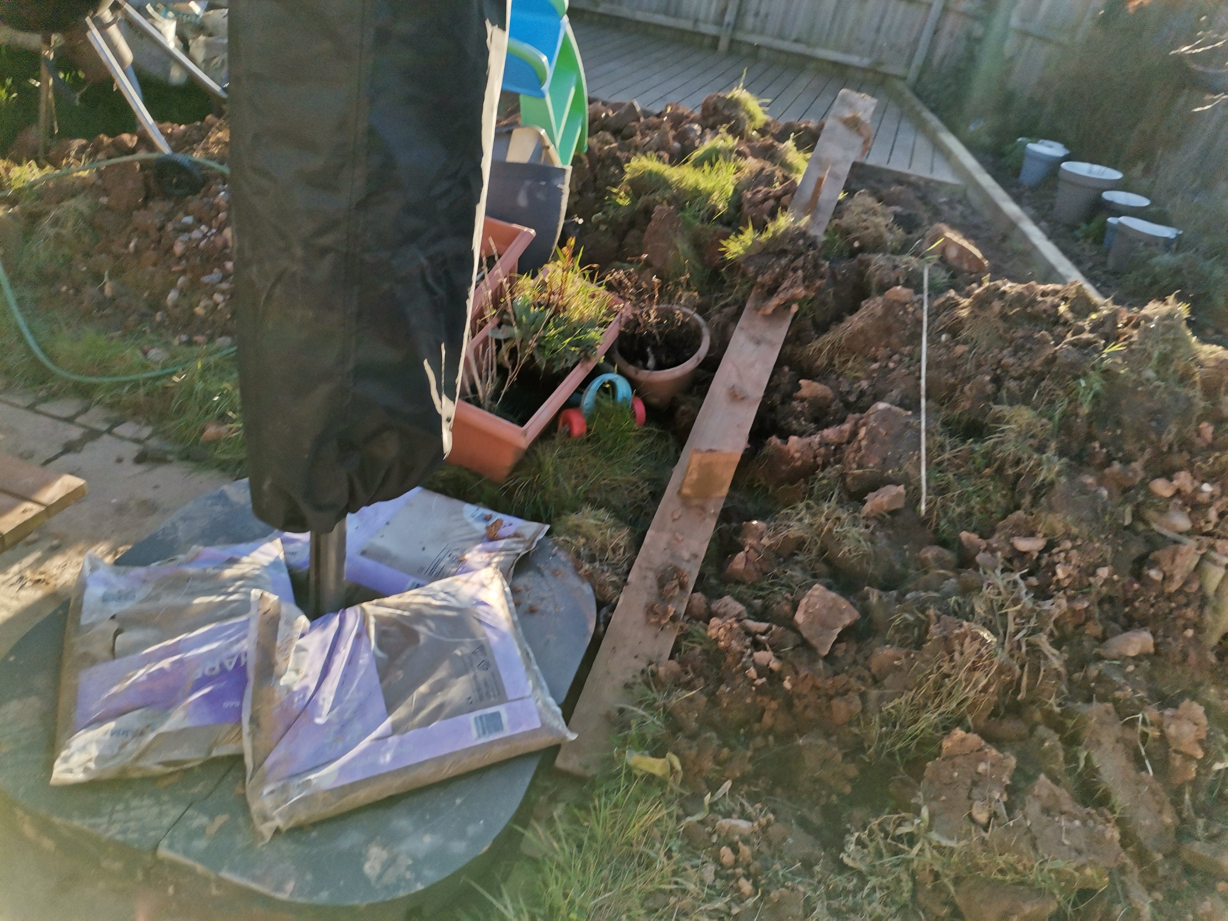 Garden area under renovation with exposed soil, garden tools, a wheelbarrow, plant pots, and bags of garden products, indicating ongoing landscaping work in a backyard during sunset.