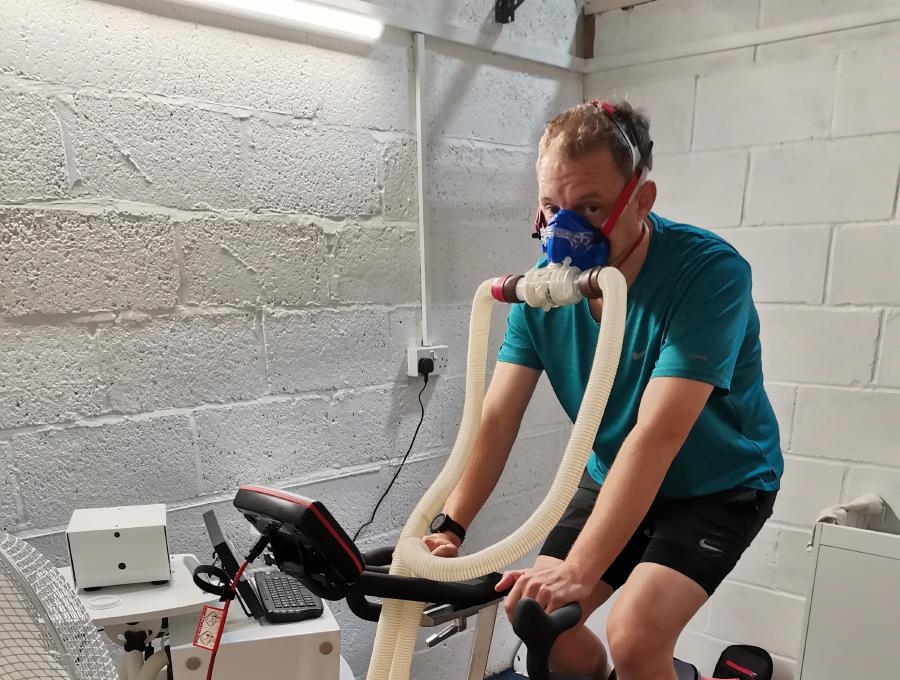 Geoffrey Hayward undergoing a cardio workout on a stationary bike inside a room with white walls, connected to a VO2 Max testing apparatus with a breathing mask, surrounded by exercise equipment.