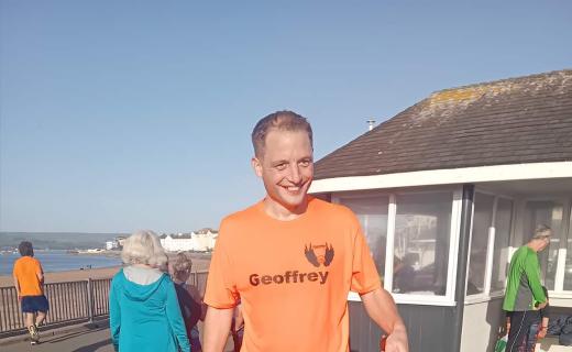 Geoffrey Hayward ringing the parkrun PB bell at Exmouth seafront with the sea in the background