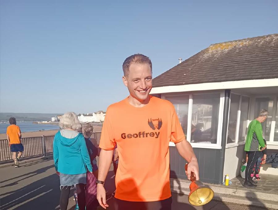 Geoffrey Hayward ringing the parkrun PB bell at Exmouth seafront with the sea in the background