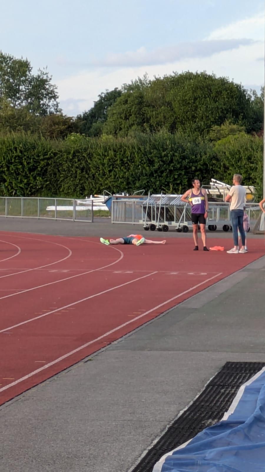 Geoffrey lying on the ground of a running track, exhausted after a race, while two other individuals stand nearby, conversing.